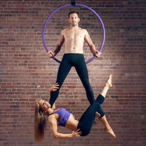 Aro aéreo X-Pole púrpura frente a una pared de ladrillos con un modelo masculino en ella y una modelo femenina colgada de sus piernas